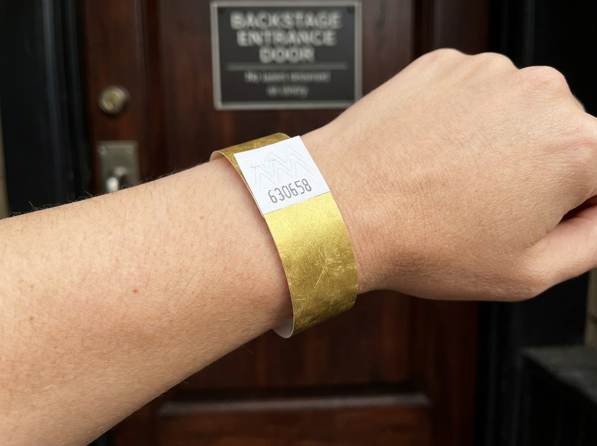 Braccialetto VIP per festival con stampa metallizzata dorata e codice a barre, indossato al polso davanti a un ingresso backstage